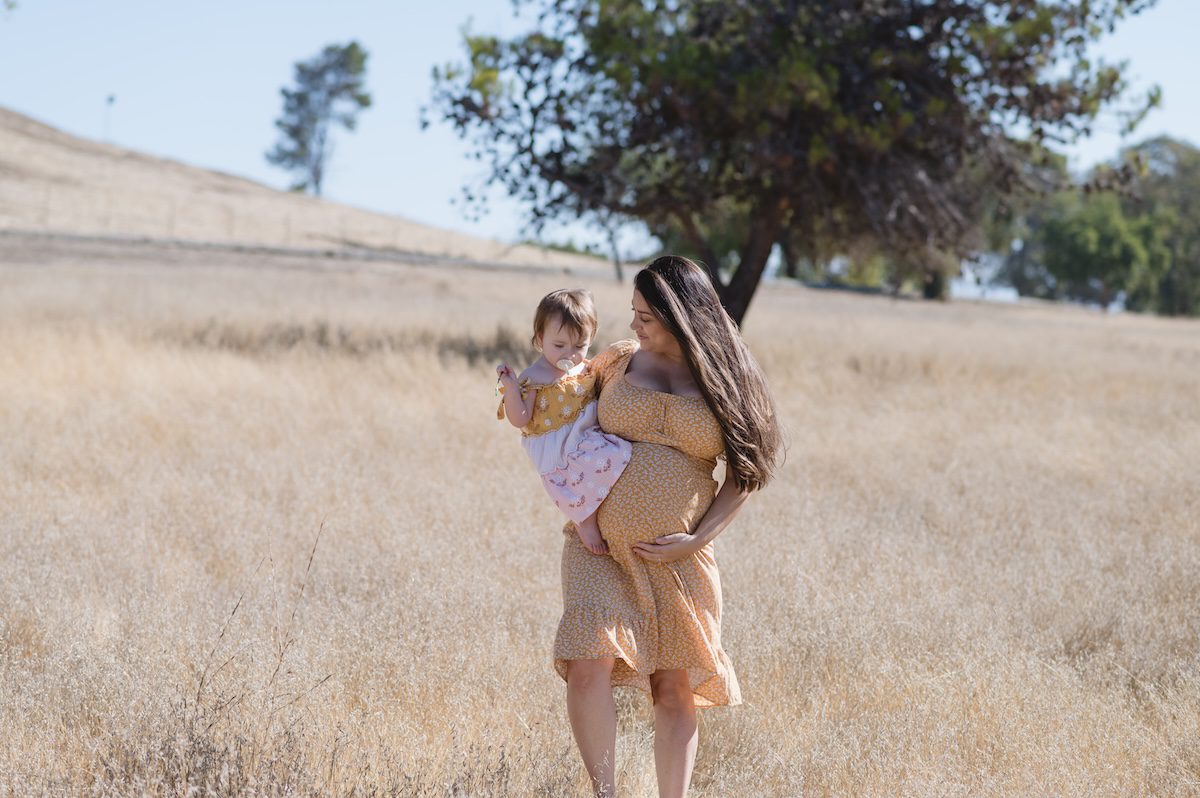 pregnant women in yellow flowy dress with a 2 year old on her hip walking in beige grass with a big tree in the background in San Jose, California by photographer Jen Vazquez