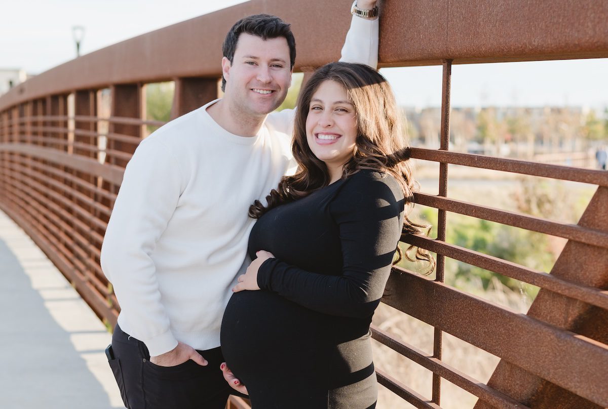 pregnant women in black long sleeve dress and the man has a white sweater and he's leaning on the a rust red bridge rails. The pregnant women is hugging her bump