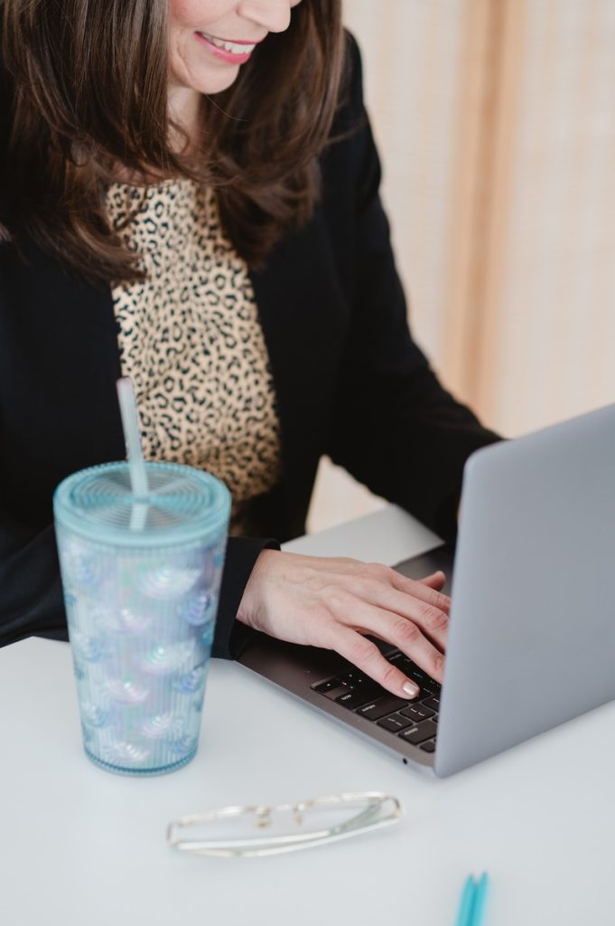 Women siting at a desk with her hands on a laptop and a plastic blue water cup with straw. The women is wearing a black suit coat and chetah shirt Reasons Your California Business Needs Brand Photography