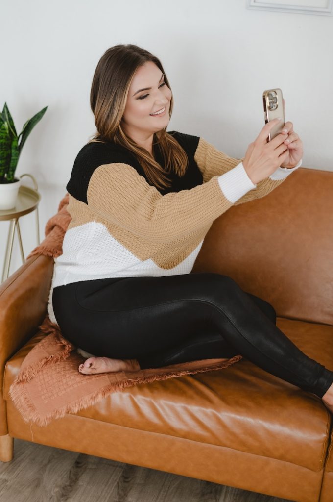 hair stylist sitting on a orange couch with a black white and beige sweater holding a cell phone up to photograph herself by Jen Vazquez Photography