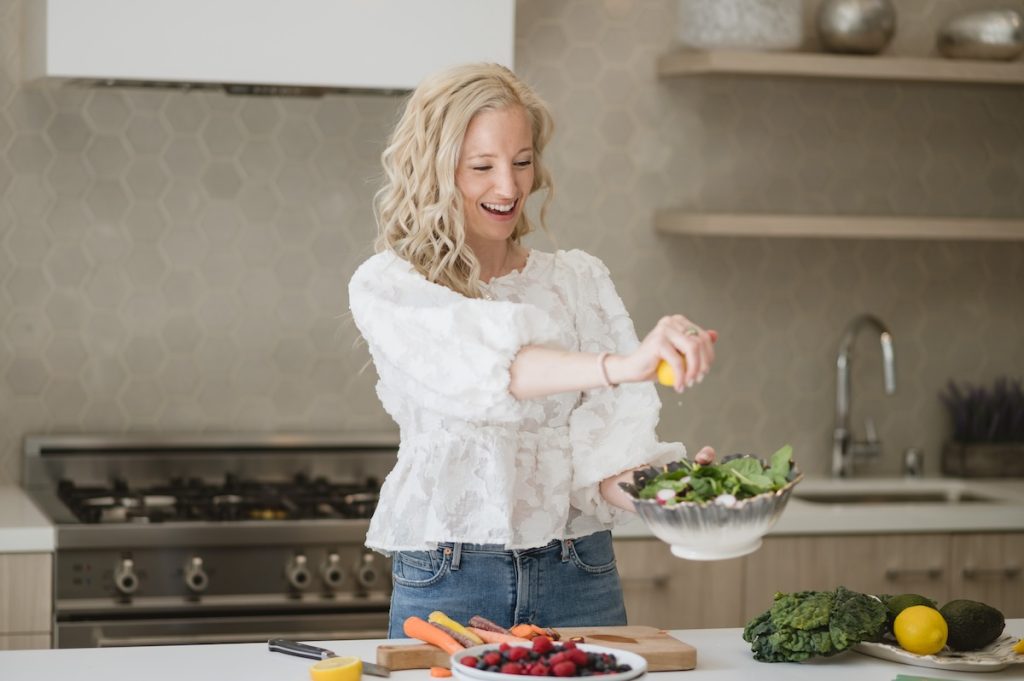 Female in a white flowy shirt with blond curled hair in a kitchen squeezing a lemon on a vibrant salad and on the counter a plate of berries, multi color carrots and ingredients to make a salad.