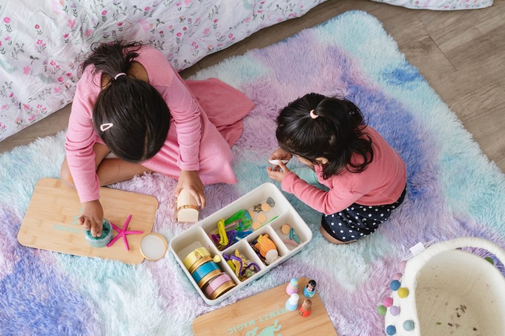 two children in pink shirts on the floor playing with a Meemzy Magic sensory kit on a multi-color rug of pinks, blues, and purple.