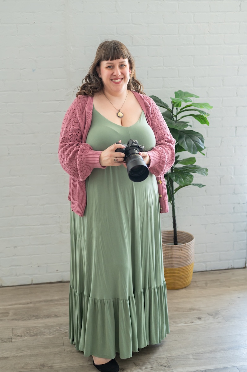 newborn photographer and business coach with green dress and pink mauve sweater holding her nikon Z6ii mirrorless camera for a photoshoot with Jen Vazquez in the Bay Area of Northern California