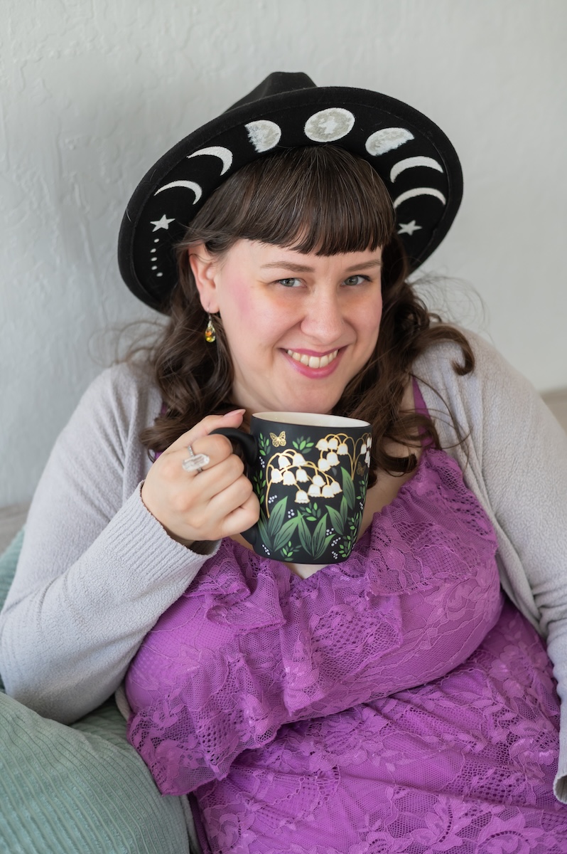 Photographer and business coach wearing a purple dress and grey sweater drinking coffee from a black coffee cup with florals for a photoshoot with Jen Vazquez in San Jose, California